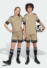 Uniformes de fútbol beiges con franjas negras, combinados con calcetines negros que tienen franjas grises. El jugador sostiene un balón de fútbol en blanco y negro.