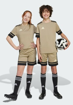 Beige voetbaluniformen met zwarte strepen, gecombineerd met zwarte sokken met grijze strepen. De speler houdt een zwart-wit voetbal vast.