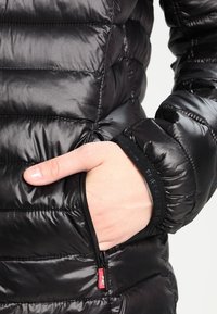 Veste matelassée noire avec un design matelassé, dotée d'une poche latérale zippée et de poignets élastiques. Le matériau est brillant et lisse.