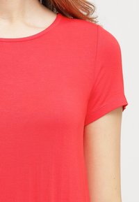 Kurzärmeliges, rundhalsiges Oberteil in leuchtendem Koralle. Hergestellt aus glattem, elastischem Stoff mit figurbetontem Schnitt und dezenten Nahtdetails an den Ärmeln.