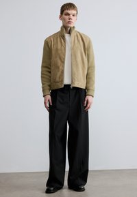 Beežist seemisnahast pommitajate jakk soonikkoes varrukatega, lukuga, kantud üle kreemika soonikkoes kampsuni ja kombineeritud lahtiste mustade pükstega.