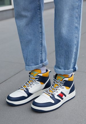Piedi con sneaker alte bianche, blu navy e gialle con calzini bianchi e jeans blu risvoltati, in piedi su un marciapiede grigio.
