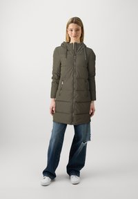 Doudoune vert olive, longueur genoux, avec capuche, motif matelassé, fermeture éclair à l'avant et deux poches zippées. Portée sur un jean déchiré.