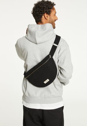Sac en bandoulière noir avec une forme arrondie, fabriqué en tissu, doté d'une fermeture éclair argentée et d'une étiquette. Porté dans le dos avec une sangle réglable.