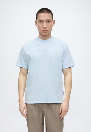 Lühikeste juustega mees, kes kannab helehalli lühikeste varrukatega T-särki ja beežide pükstega, seisab tavalise valge tausta ees.