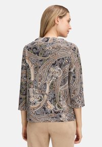 Kobieta stojąca bokiem, prezentująca tył bluzki o wzorze paisley, z rękawami do trzech czwarte, w beżowych, czarnych i zielonych detalach, w połączeniu z beżowymi spodniami.