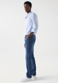 Svijetloplavi prugasti košulja dugih rukava, plave traperice, bijeli tenisice; glatka tkanina, opušteni kroj, bočni pogled, s rukama u džepovima.