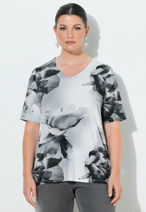 Kvinde med mørkt hår i en elegant knold bærer perleclusterøreringe og en hvid V-hals skjorte med store sorte og grå blomsterprint.