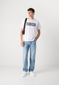 Man wearing white Calvin Klein t-shirt, light blue jeans, white sneakers, and black crossbody bag, standing against plain white background.
