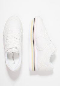 Baskets blanches à lacets avec une semelle intermédiaire rayée pastel jaune, rose et noire, vues de dessus et de côté sur un fond blanc.