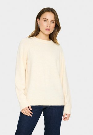 Donna che indossa un maglione a maniche lunghe color crema e jeans blu scuro, in piedi contro uno sfondo bianco semplice.