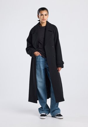 Zwarte lange jas met een dubbelzijdig ontwerp, brede revers en opgerolde mouwen, gecombineerd met lichtblauwe brede jeans en zwarte sneakers.