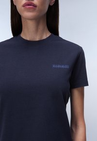 Námořnická modrá bavlněná tričko s kulatým výstřihem a krátkými rukávy; na levé straně hrudi se nachází malé, světle modré logo značky.
