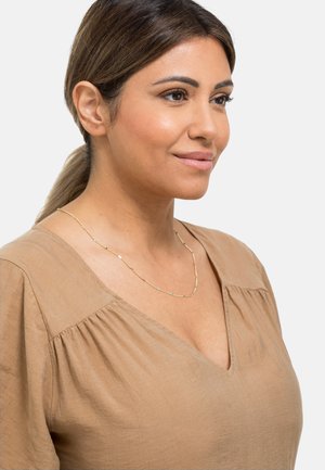 Collier doré avec des maillons allongés et texturés. Porté avec un haut beige présentant un col en V et des détails de manches froncées.