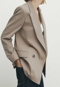 Blazer beige à double boutonnage avec des revers nets, deux boutons et des épaules structurées. Dispose de poches latérales et d'une texture de tissu lisse.