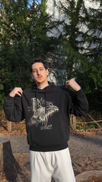 Muž vonku v čiernej mikine s kapucňou s veľkým motýľom a nápisom "NOCTURNAL SPIRIT," stojaci pred vysokými zelenými stromami a spevnenou cestou.