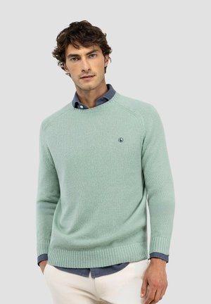 Hombre con jersey de cuello redondo verde claro sobre camisa azul de botones, con las manos en los bolsillos, sobre fondo gris liso.