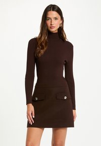 Maglione a collo alto marrone a costine abbinato a una gonna a pieghe. La gonna presenta due tasche anteriori con bottoni metallici circolari.