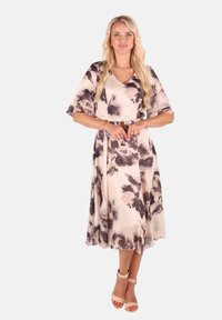 Blumenkleid in sanftem Beige mit großen grauen und lila Mustern, V-Ausschnitt, ausladenden Ärmeln und einem fließenden A-Linien-Rock, kombiniert mit nude-farbenen Absätzen.