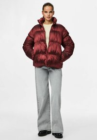 Burgundi pufinė striukė su blizgiu paviršiumi, aukštu apykakle, užtrauktuku priekyje ir siuvinėto dizaino. Suderinta su pilkų plačių džinsų.