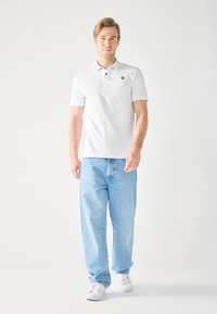 Chemise polo blanche avec un logo noir, associée à un jean bleu clair ample et des baskets blanches, présentée sur un fond neutre.