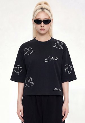 Jeune femme aux longs cheveux blonds en queue de cheval, portant des lunettes de soleil noires et un t-shirt noir avec un oiseau blanc et des motifs et textes "Liberté".