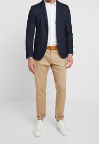 Marinblazer med texturerad tyg, vit skjorta med knappar, khakifärgade chinos, brun bälte och vita sneakers. Upprullade ärmar på byxorna.