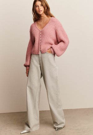 Woman wearing a pink fuzzy buttoned cardigan, light gray wide-leg jeans, and metallic pointed-toe shoes, standing against a plain background.