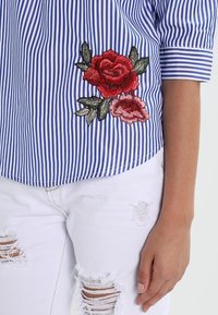 Camicetta a righe blu e bianche con rose rosse ricamate e foglie verdi sul lato sinistro. Indossata con jeans bianchi strappati.