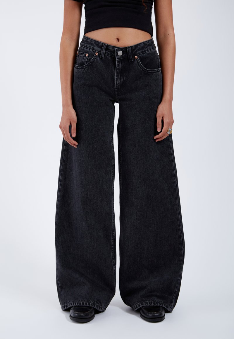 Vrouw die hoge taille, brede zwarte jeans draagt, zwart cropped top, zwarte schoenen en een ring aan haar rechterhand, staand op een witte achtergrond.