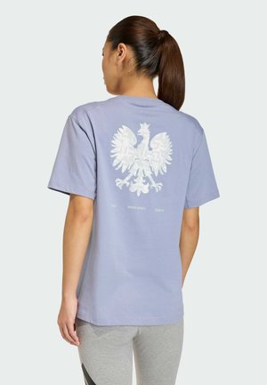 Kobieta z brązowym kucykiem, ubrana w jasnofioletową koszulkę z dużym białym orłem oraz napisem "2025 Winter Sports Team PL" na plecach.