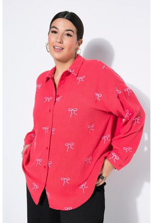 Vrouw met donker haar in een losse rode blouse met knopen, bedrukt met kleine witte strikjes, staand met handen in de zakken tegen een effen achtergrond.