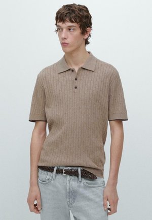 Giovane uomo che indossa una polo beige a maniche corte con lavorazione a trecce infilata in jeans grigio chiaro con cintura intrecciata marrone, che guarda a sinistra.