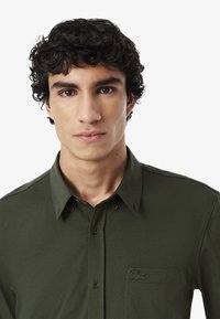 Camicia verde oliva con bottoni realizzata in tessuto liscio, dotata di taschino sul petto e dettagli del logo ricamato. Collo classico e chiusura con bottoni.