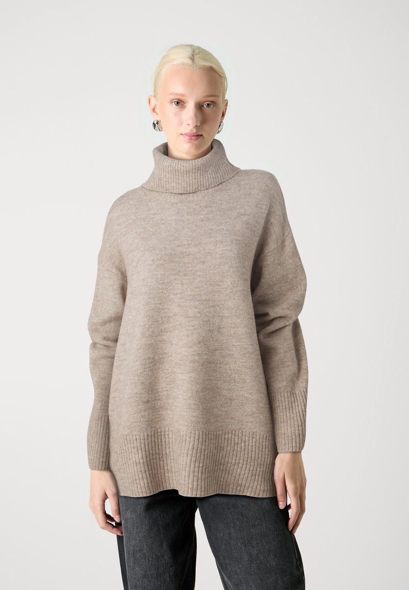 Beige Rollkragenpullover mit gerippten Bündchen und Saum; weiche Textur, lockere Passform. Kombiniert mit dunklen Jeans steht das Modell vor einem schlichten Hintergrund.