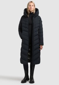 Abrigo puffer largo negro con capucha, con patrones acolchados y cremallera completa. Llevado sobre un conjunto negro y combinado con botas negras.
