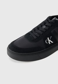 Černé nízké tenisky s černými tkaničkami, semišovými detaily, silnou gumovou podrážkou a bílým logem "cK" na bočním panelu.