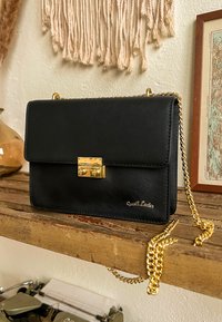 Schwarze Lederhandtasche mit einer goldenen Kettenriemen und einem Druckverschluss. Sie verfügt über eine glatte Textur und einen geprägten Markennamen auf der Vorderseite. Rechteckige Form.