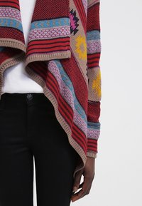Cardigan tricoté coloré avec des motifs géométriques rouges, bleus, roses et jaunes, porté sur un haut blanc et un pantalon noir, main détendue sur le côté.