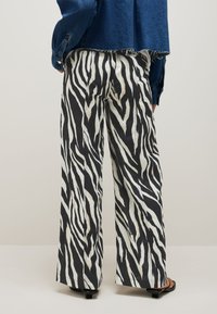 Woman wearing wide-leg pants with black and white zebra stripes, a cropped denim jacket, and black heeled sandals, standing on a white floor.