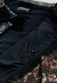 Zwarte stoffen kledingstuk met een fluwelen strik, een metalen knoop en een paillettenpatroon in brons en goud. Glad textuur met contrasterende details.
