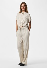 Giovane donna con le mani in tasca, indossa una camicia beige a righe sottili con maniche corte annodata e pantaloni ampi coordinati con scarpe nere.