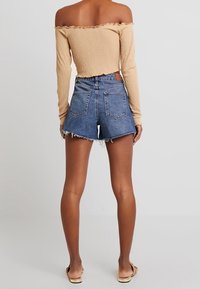 Beige texturovaný top s odhalenými rameny a třásněmi na lemování, který je kombinován s modrými riflovými šortkami s vysokým pasem, jež mají klasický střih a distressed detaily.