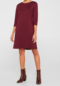 Burgunderfarbenes Kleid mit Dreiviertelärmeln, A-Linien-Design, aus glattem Stoff, kombiniert mit braunen Leder-Stiefeletten und einer strukturierten Oberfläche.