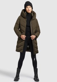 Olijfgroen pufferjack met een capuchon, met chevronstiksel en een rits aan de voorkant. Draagt over zwarte leggings en enkellaarsjes.