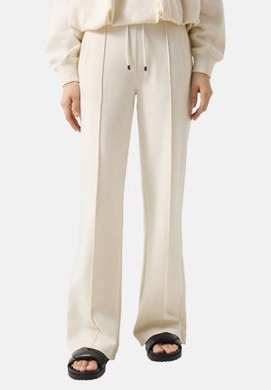 Person iført cremekulørte bukser med vide ben og forreste plisseringer, kombineret med sorte sandaler med åben tå, stående mod en ensfarvet baggrund.