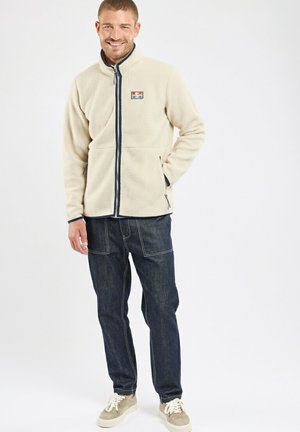 Homme souriant, portant une veste polaire beige à fermeture éclair, un jean bleu foncé et des baskets beiges, debout devant un fond blanc.