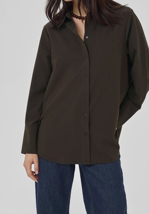 Kobieta ubrana w ciemnobrązową koszulę w cienkie prążki na guziki i niebieskie dżinsy, z jedną ręką w kieszeni i widocznym koralikowym naszyjnikiem.