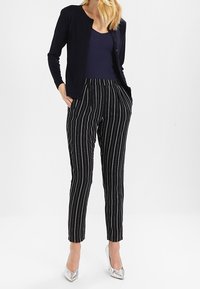 Schwarz-weiße gestreifte Hose mit hohem Bund, kombiniert mit einem marineblauen Cardigan und einem Tanktop, ergänzt durch silberne High Heels.