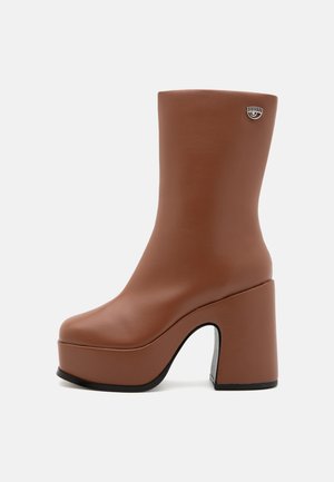 Bottines en simili cuir marron avec un large talon bloc, bout arrondi et un détail argenté près du haut. Texture lisse et design épuré.